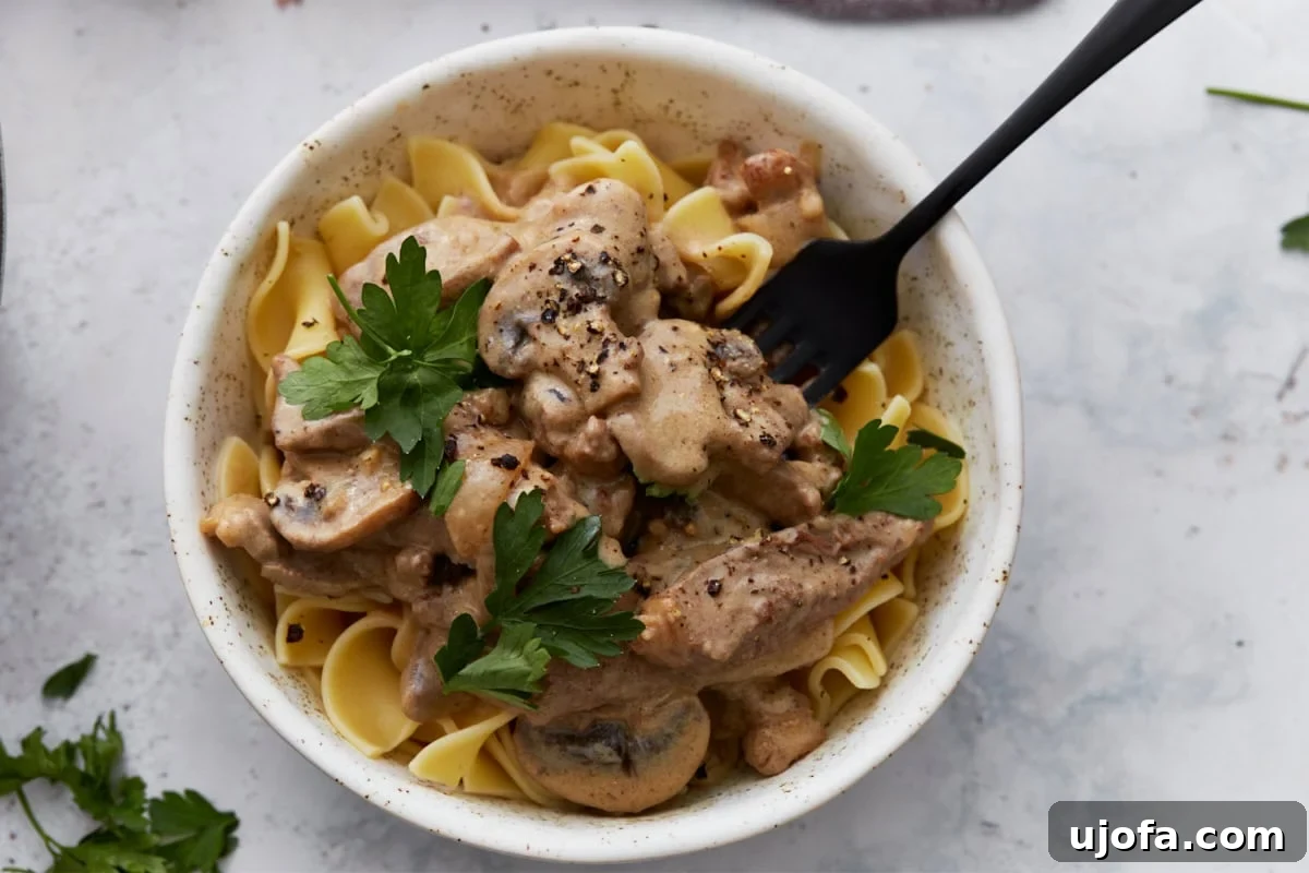 Close-up of a serving of gluten-free beef stroganoff spooned over egg noodles in a white bowl, topped with fresh parsley and cracked black pepper, with a black fork tucked in.