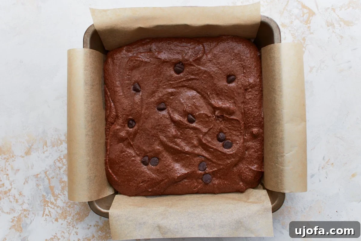 Raw keto brownie batter spread evenly in a parchment-lined square pan, topped with extra chocolate chips.