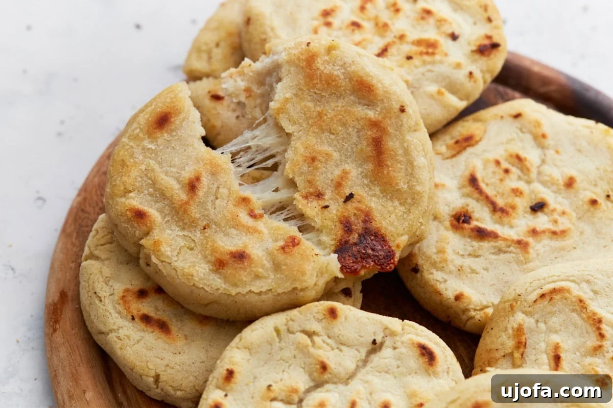 Savory Salvadoran Pockets 13 Close-up of a cheesy pupusa torn in half, showing melted cheese stretching from the center. The pupusa has a golden, crispy exterior and sits on a wooden plate with other freshly cooked pupusas