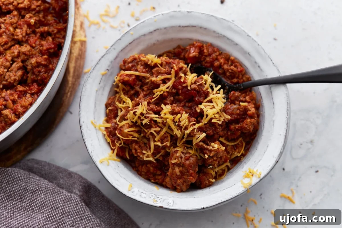 Hearty Keto Chili 10 A white bowl of keto chili topped with shredded cheddar cheese, with a black spoon ready for serving.