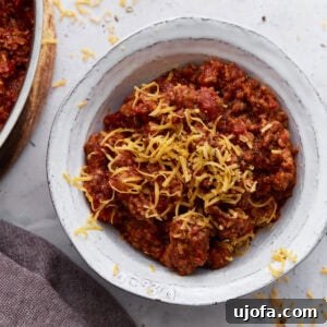A white bowl filled with keto chili topped with shredded cheddar cheese, placed on a light surface with cheese scattered around.