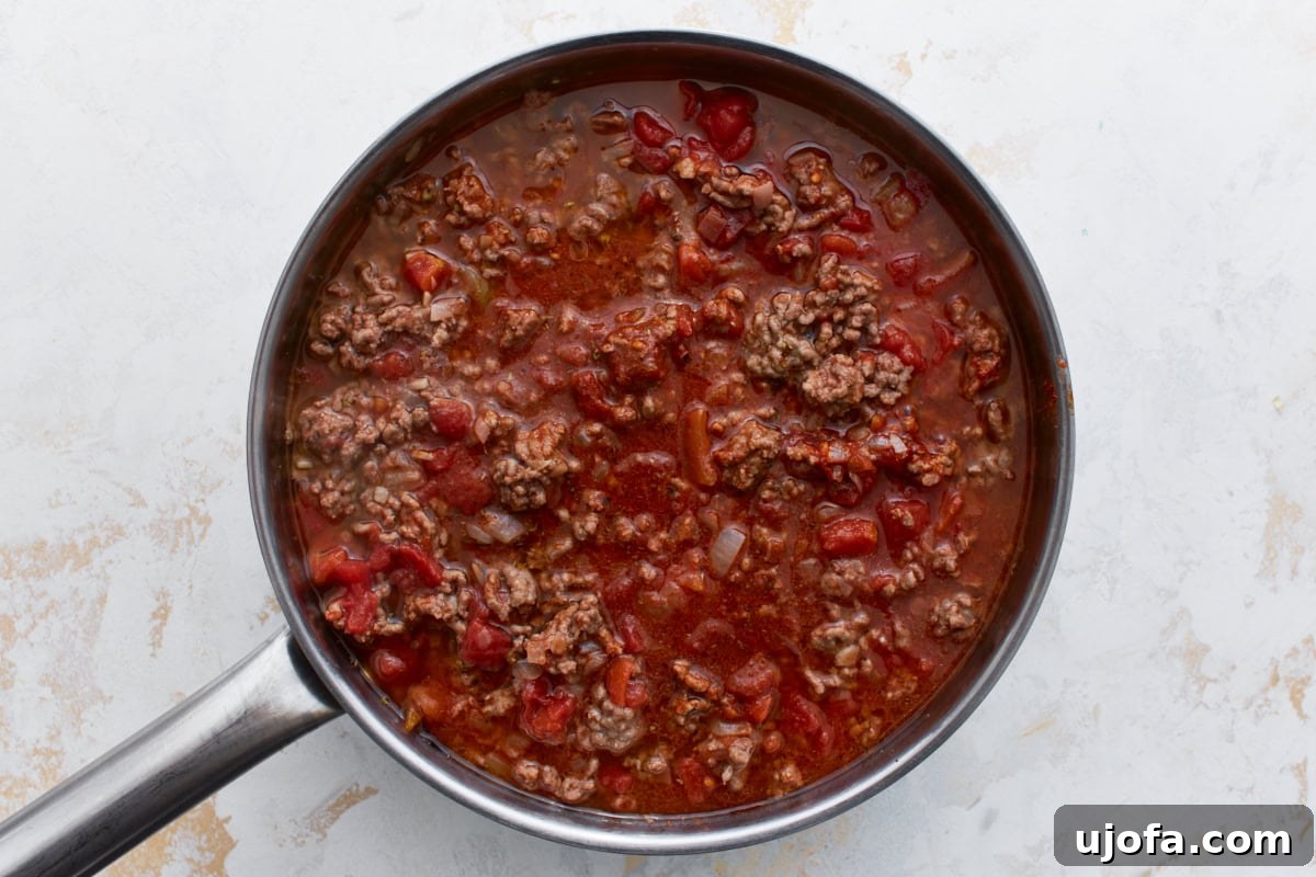 Hearty Keto Chili 7 Ground beef, diced tomatoes, and broth simmering together in a skillet with visible liquid and rich red color.