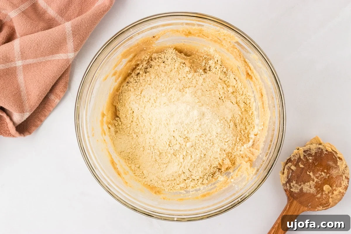 Chickpea flour being gently poured into the wet mixture in a glass mixing bowl, signaling the start of combining dry and wet elements.