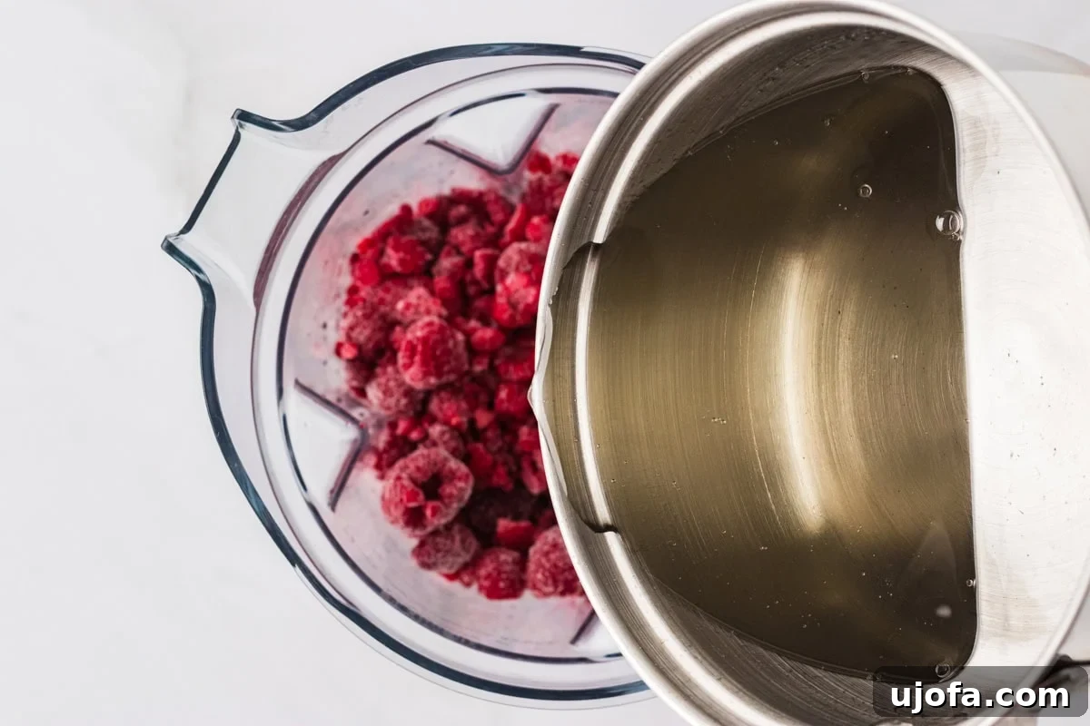 A saucepan of freshly made simple syrup being poured into a blender filled with frozen raspberries.