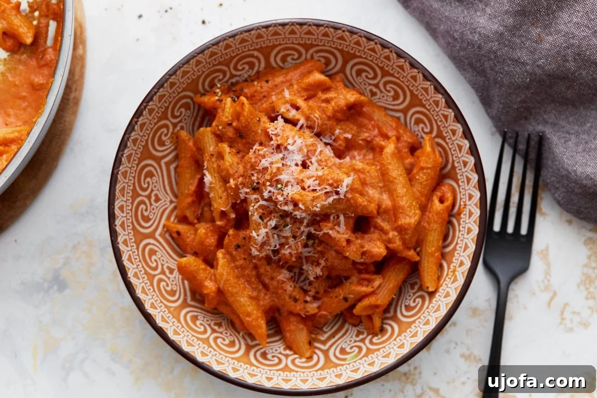 A serving of gluten-free penne pasta coated in creamy vegan vodka sauce, topped with grated vegan parmesan and cracked black pepper in a patterned bowl.