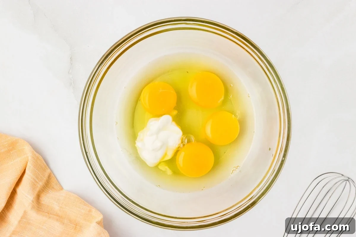 Guilt-Free Golden Cornbread 4 Mixing bowl with four cracked eggs and sour cream before whisking, sitting on a white counter with a whisk to the side. Ready for wet ingredients to be combined.