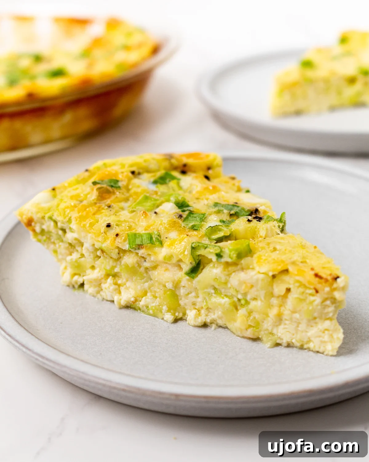Fluffy Zucchini Egg Bake 2 A vibrant slice of crustless zucchini quiche, generously topped with fresh green onions, resting on a sophisticated gray plate. The full, perfectly baked quiche is beautifully blurred in the background, hinting at its deliciousness.