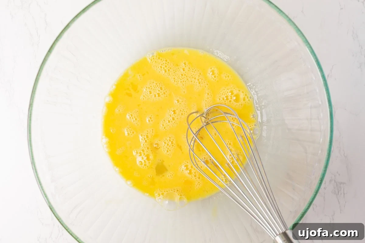 Fluffy Zucchini Egg Bake 6 A shiny metal whisk partially submerged in a bowl of perfectly whisked eggs, showcasing their smooth, uniform texture, awaiting further ingredients for the quiche mixture.