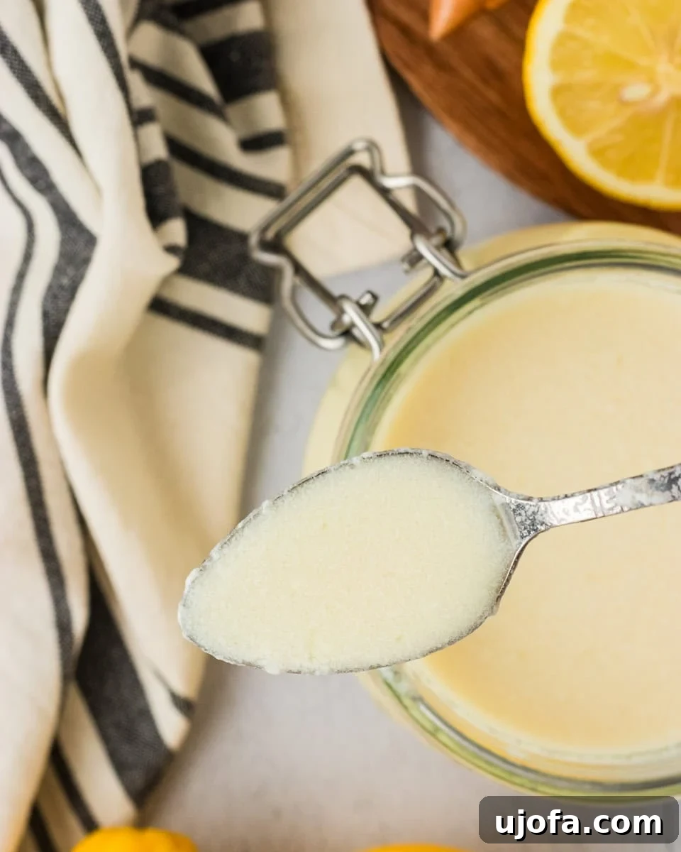 A spoonful of freshly made vegan buttermilk held above a glass jar, showing its creamy texture with lemons and a striped kitchen towel in the background.