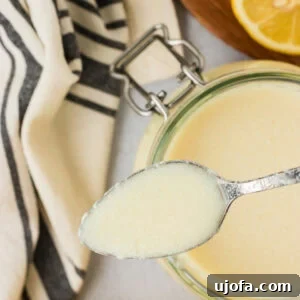 A spoonful of freshly made vegan buttermilk held above a glass jar, showing its creamy texture with lemons and a striped kitchen towel in the background.