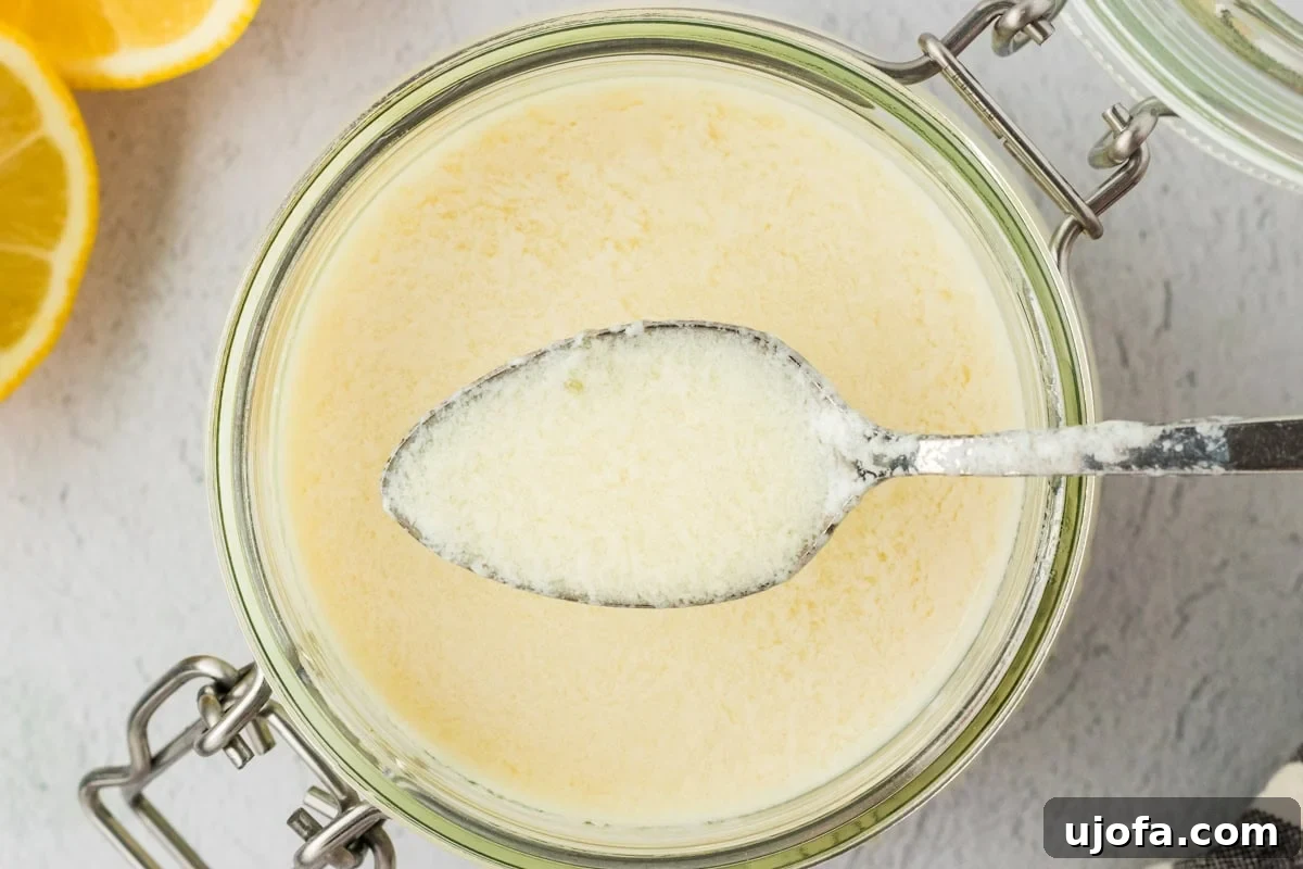 A close-up of a spoon dipped into a jar of vegan buttermilk, showing its lightly curdled, creamy consistency with sliced lemons in the background.