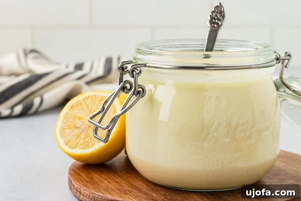 A glass jar filled with creamy vegan buttermilk sits on a wooden board with a spoon inside, next to a halved lemon and a striped kitchen towel in the background.