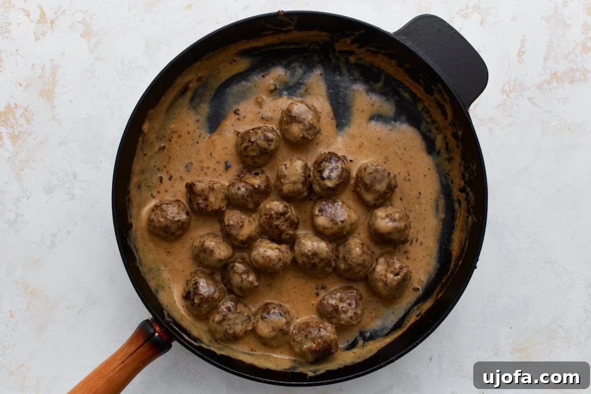 The browned Swedish meatballs submerged and simmering in the creamy brown sauce within the skillet, absorbing its rich flavors.