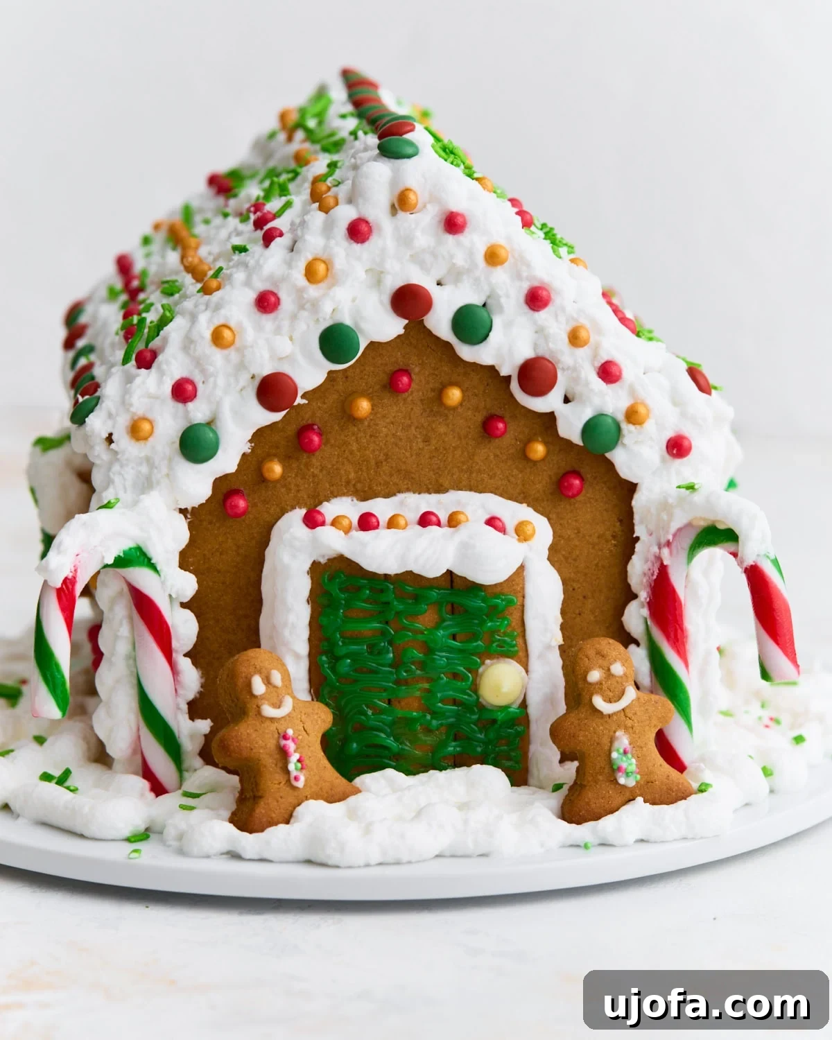 A fully assembled gluten-free gingerbread house decorated with white royal icing, colorful candies, candy canes, and two small gingerbread people by the doorway.