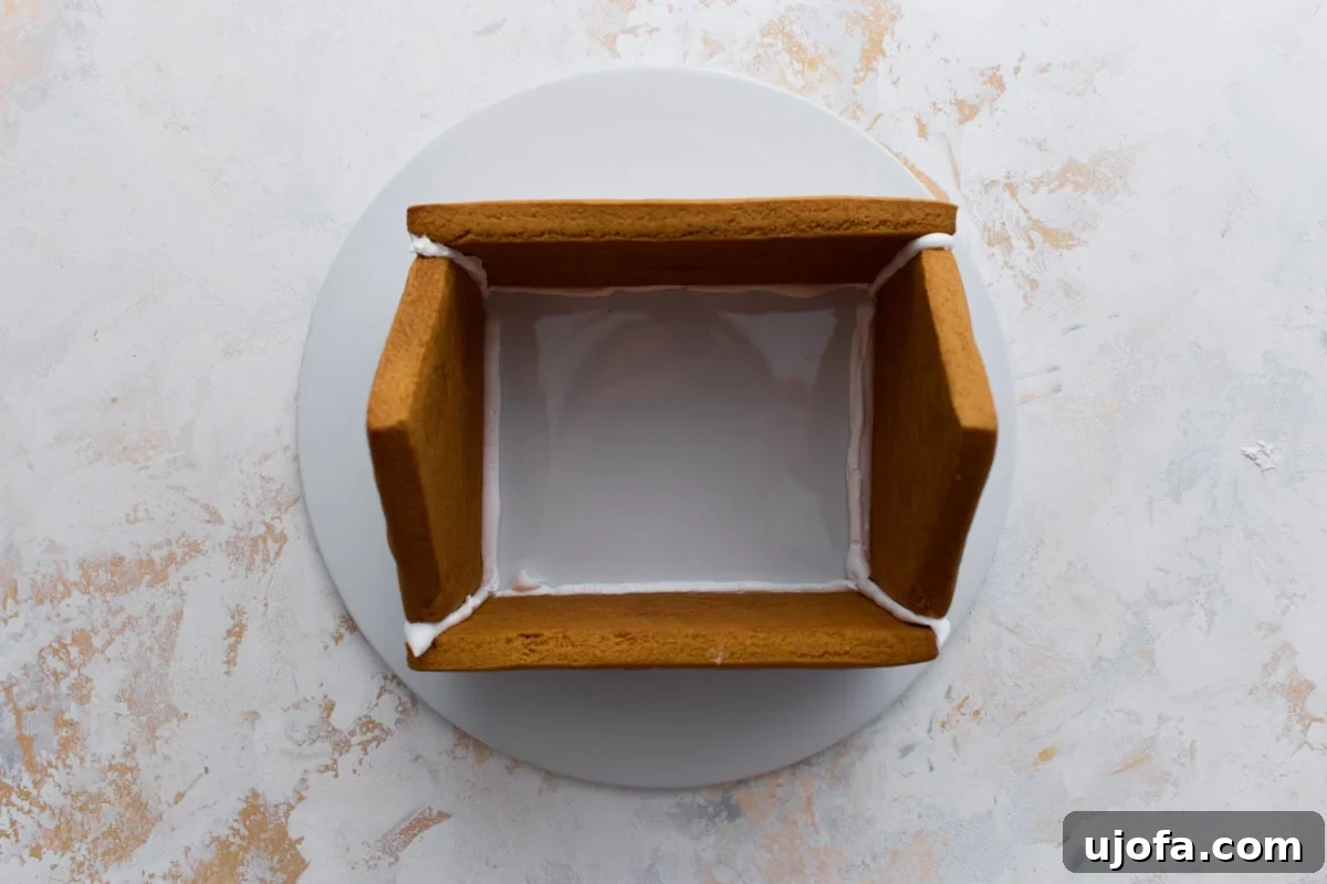 The base of the gingerbread house being assembled on a round platter, with four cookie walls secured together using white royal icing.