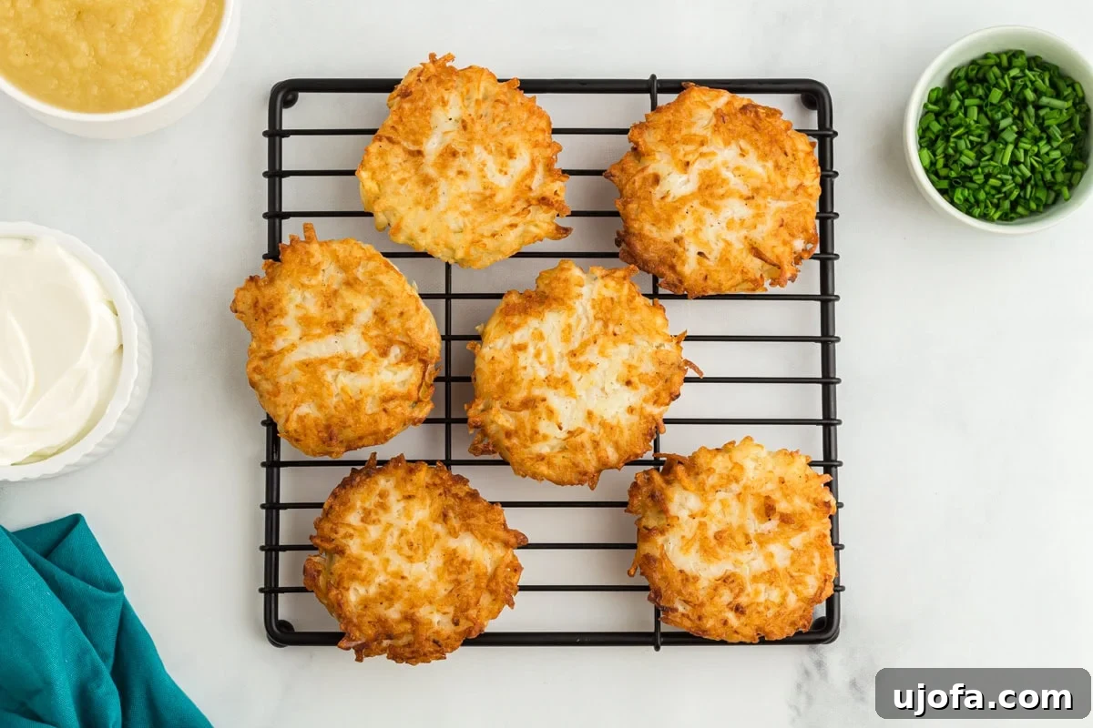 A cooling rack laden with six freshly fried, golden-crisp gluten-free potato latkes, with small bowls of applesauce, sour cream, and chopped chives arranged artfully for serving.