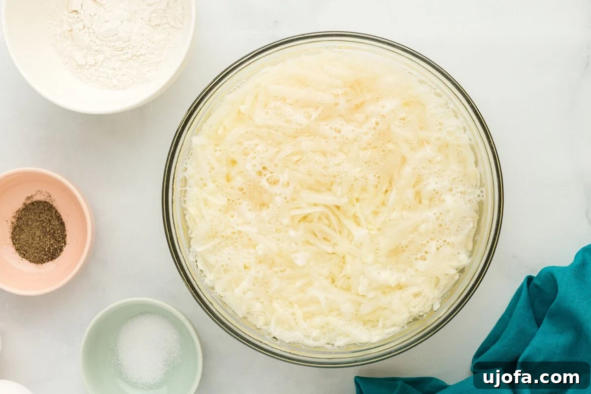 Grated russet potatoes soaking in a clear glass bowl filled with cold water, illustrating the essential step to remove excess starch for crispier latkes.