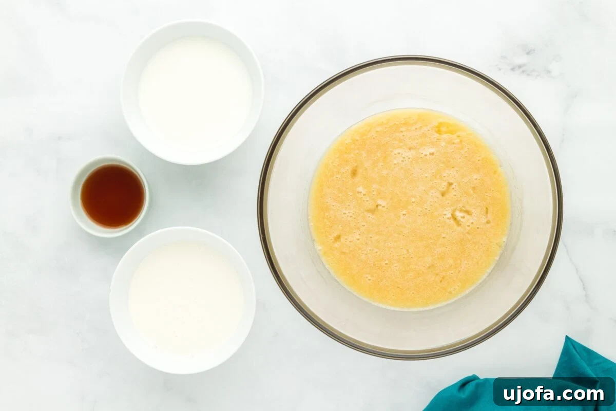 Velvet Coconut Dream 5 A large glass bowl contains a creamy, whisked mixture of sugar, butter, and eggs. Surrounding the bowl are smaller bowls holding milk, heavy cream, and vanilla extract, all set on a light wooden surface.
