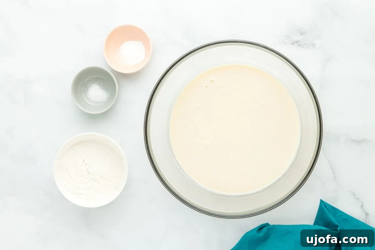 Velvet Coconut Dream 6 A smooth, light yellow batter fills a clear glass mixing bowl. Around the bowl, small white dishes hold gluten-free flour, baking powder, and a pinch of salt, neatly arranged on a light wooden surface.