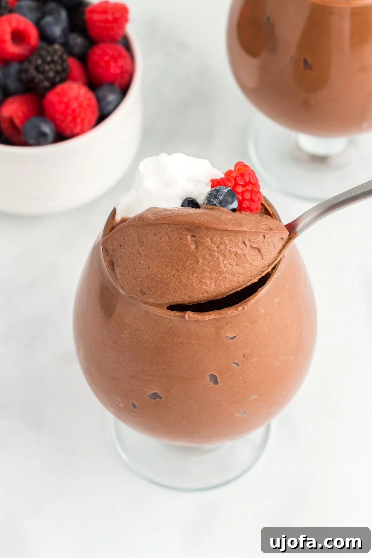 Decadent Vegan Chocolate Treat 2 A glass filled with vegan chocolate mousse topped with whipped coconut cream and fresh berries, with a spoon scooping into the mousse and a bowl of mixed berries in the background.