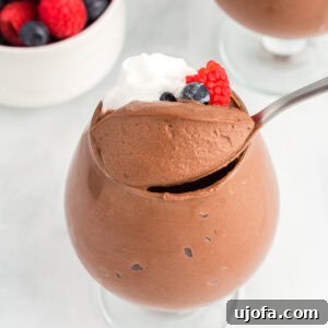 Decadent Vegan Chocolate Treat 3 A glass filled with vegan chocolate mousse topped with whipped coconut cream and fresh berries, with a spoon scooping into the mousse and a bowl of mixed berries in the background.