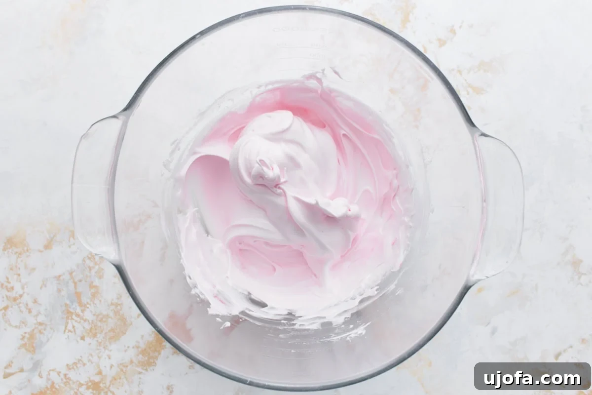 Light pink tinted meringue in a glass bowl after vanilla extract and gel food coloring have been mixed in.