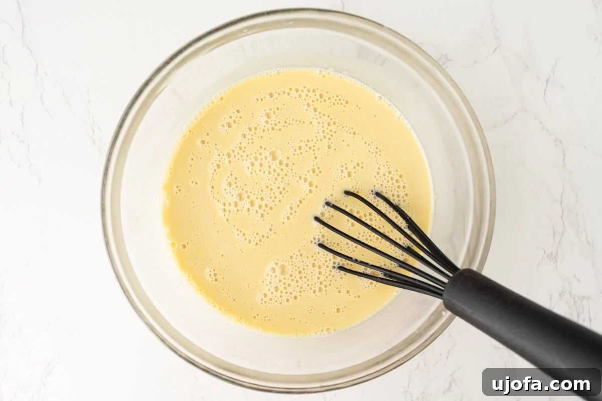 Plant Based Crème Brûlée Bliss 8 A stream of warm coconut milk mixture being slowly poured into whisked egg yolks in a glass bowl, creating a silky, combined custard base.