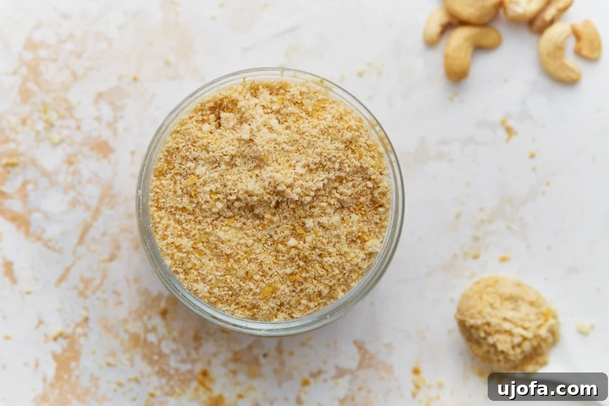 Plant Based Parmesan Perfection 7 A top-down view of a clear glass jar filled with crumbly vegan parmesan cheese, with raw cashews scattered around its base on a light surface.