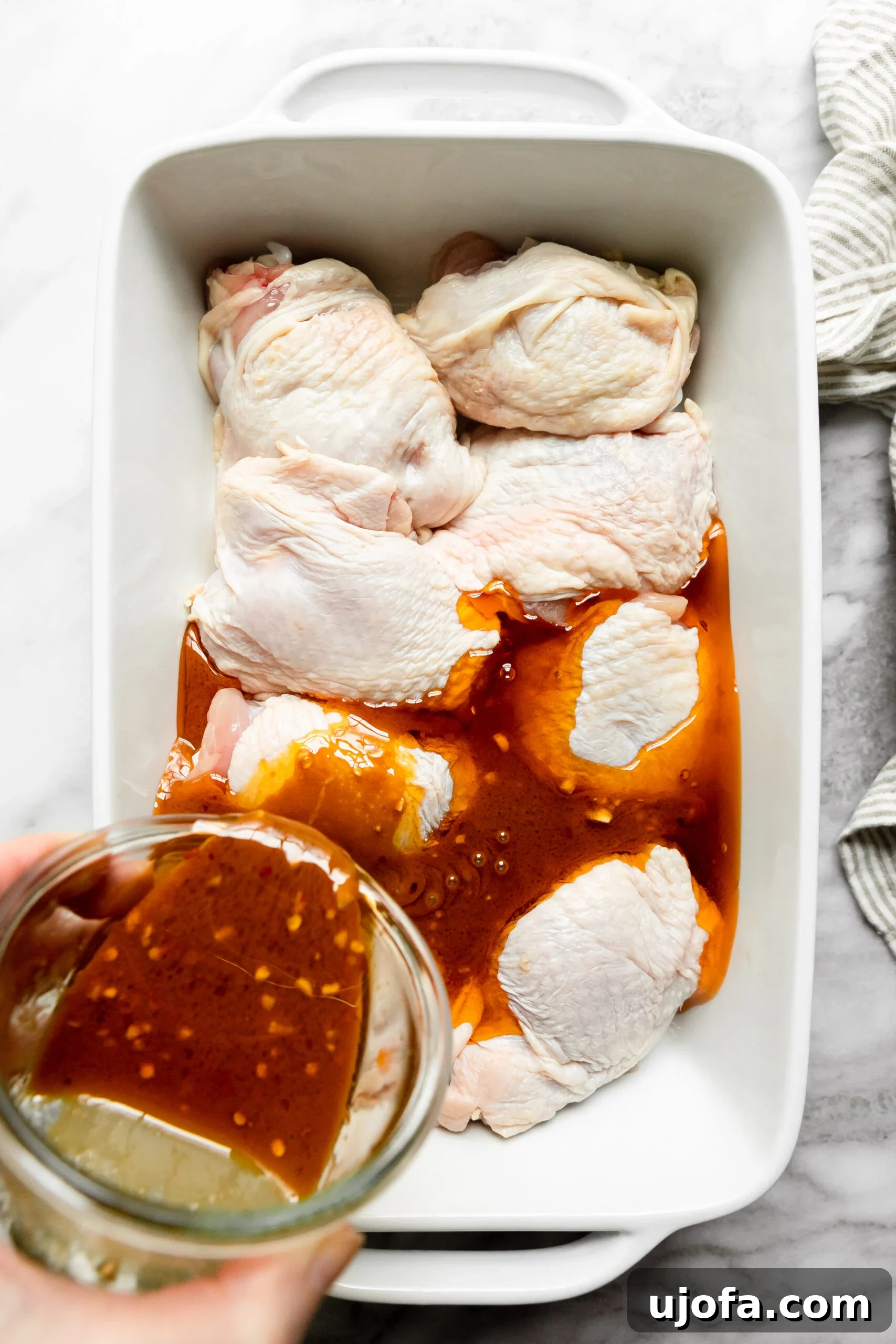 Juicy Keto Chicken Thighs with a Healthy Zesty Marinade 5 A white casserole dish lined with raw chicken thighs, with a glass bowl of the flavorful marinade being poured over them, preparing for marination.