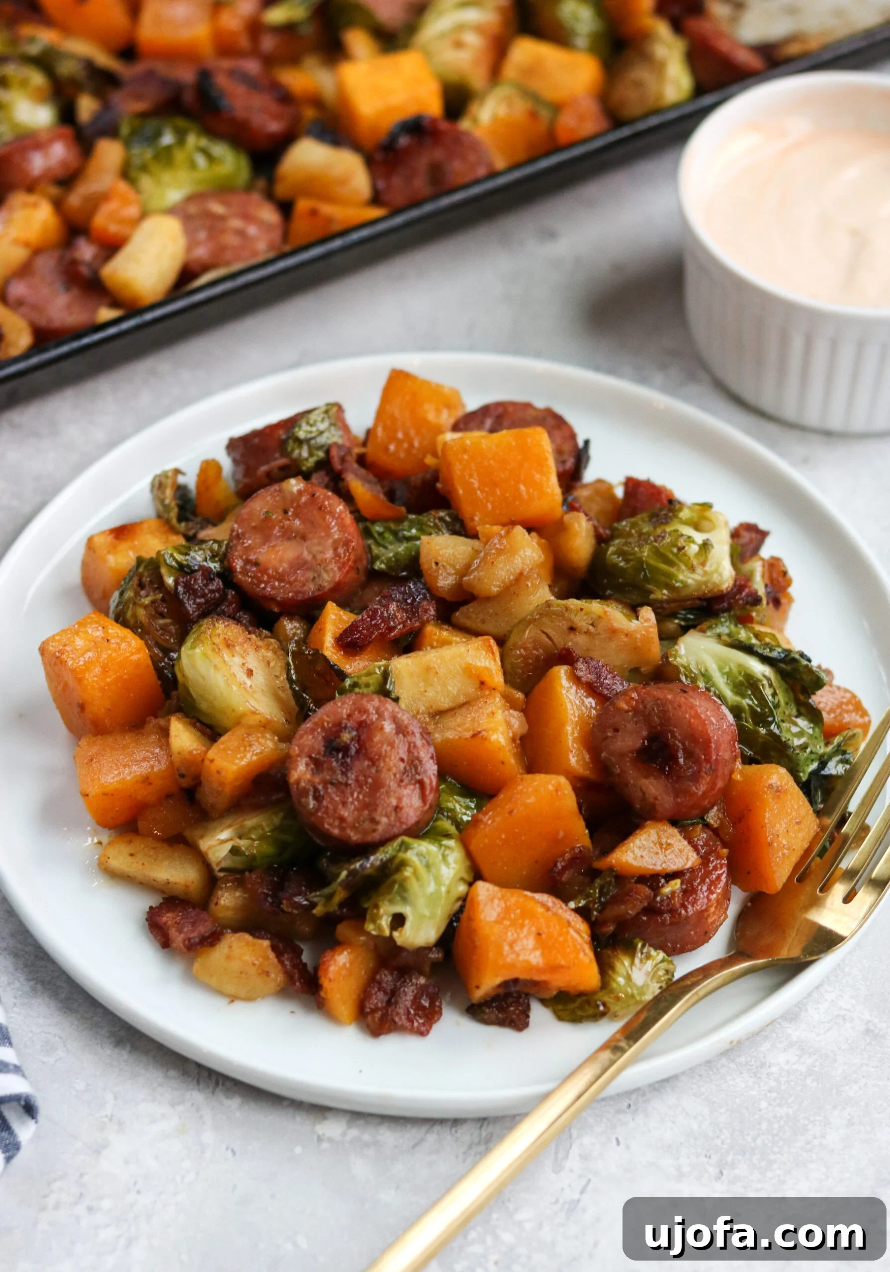 Close-up of roasted chicken sausage, butternut squash, Brussels sprouts, apples, and bacon on a sheet pan, showcasing their caramelized textures.