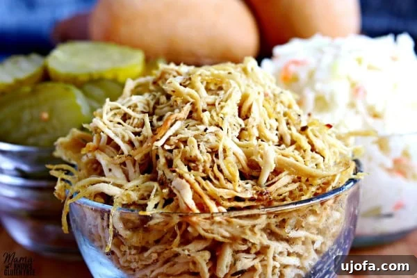 A spread of rotisserie style shredded chicken in a bowl, bread and butter pickles, creamy coleslaw, and gluten-free ciabatta rolls, ready for making sandwiches.