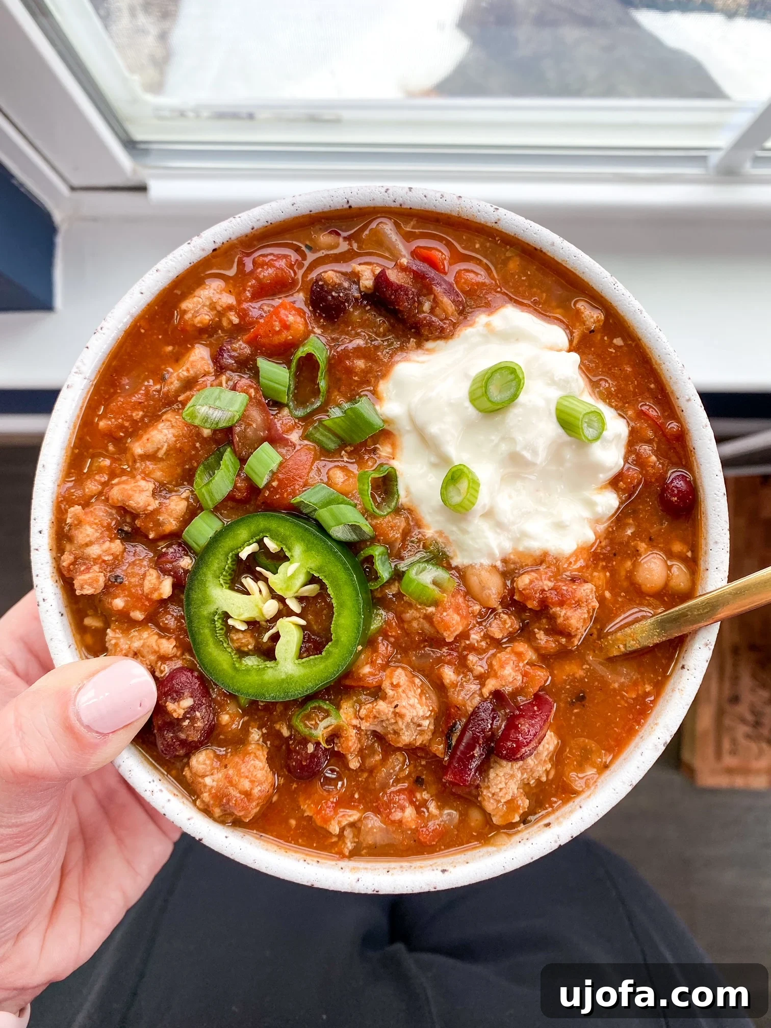 A person holding a bowl of steaming slow cooker turkey chili, topped with sour cream and a sprig of cilantro, ready to be enjoyed.