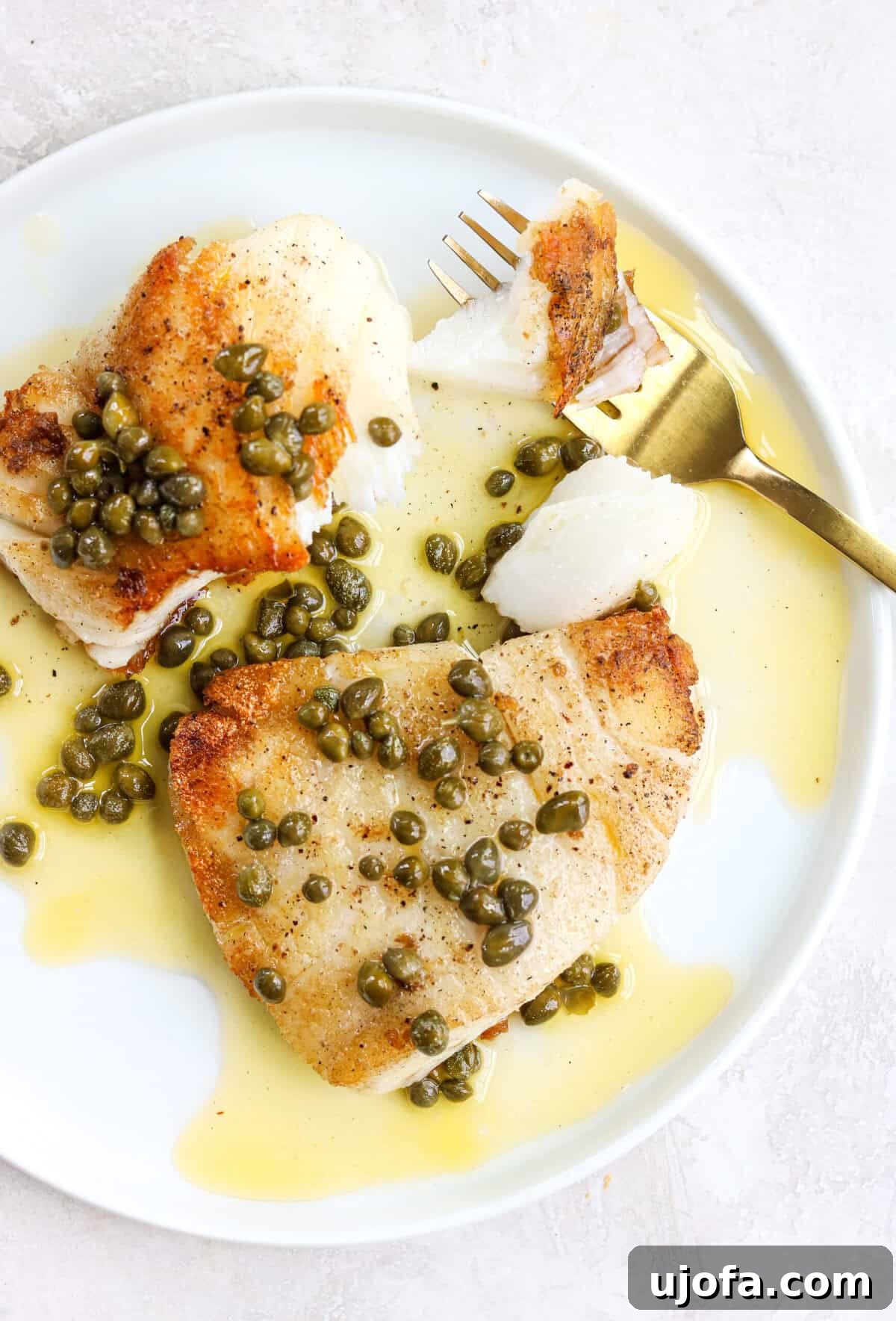 20-Minute Chilean Sea Bass with Bright Lemon Caper Butter 2 Crispy pan-seared white fish on a white plate with lemon caper sauce and a gold fork with a bit of fish on it, showcasing a perfect crust and flaky interior.
