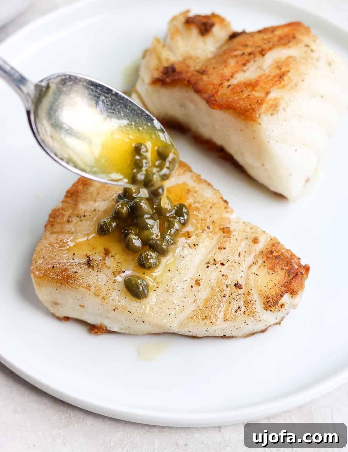 20-Minute Chilean Sea Bass with Bright Lemon Caper Butter 9 A silver spoon elegantly drizzling bright lemon caper butter over a perfectly cooked piece of Chilean sea bass on a white plate, ready for serving.
