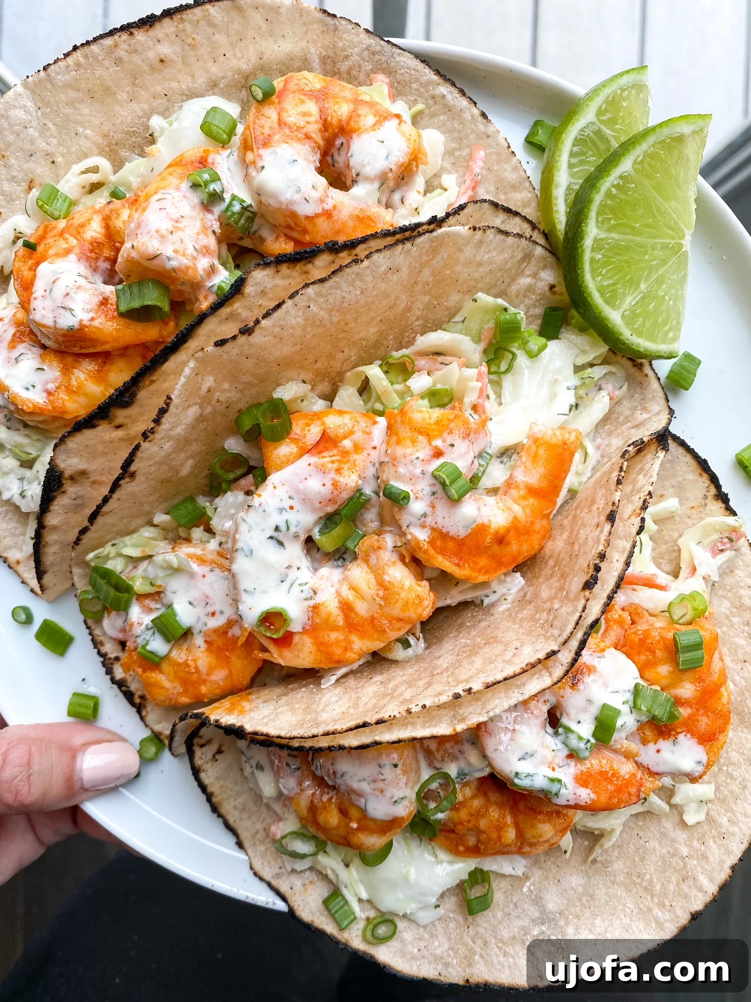 A hand holds a white plate showcasing a trio of vibrant Paleo Buffalo Shrimp Tacos, garnished with fresh herbs and a lime wedge, ready to be served.