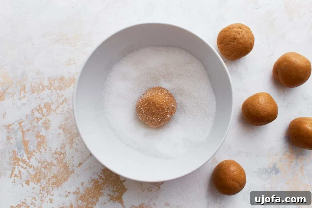 A gluten-free peanut butter dough ball being rolled in granulated sugar inside a white bowl, with additional cookie balls waiting on the side.