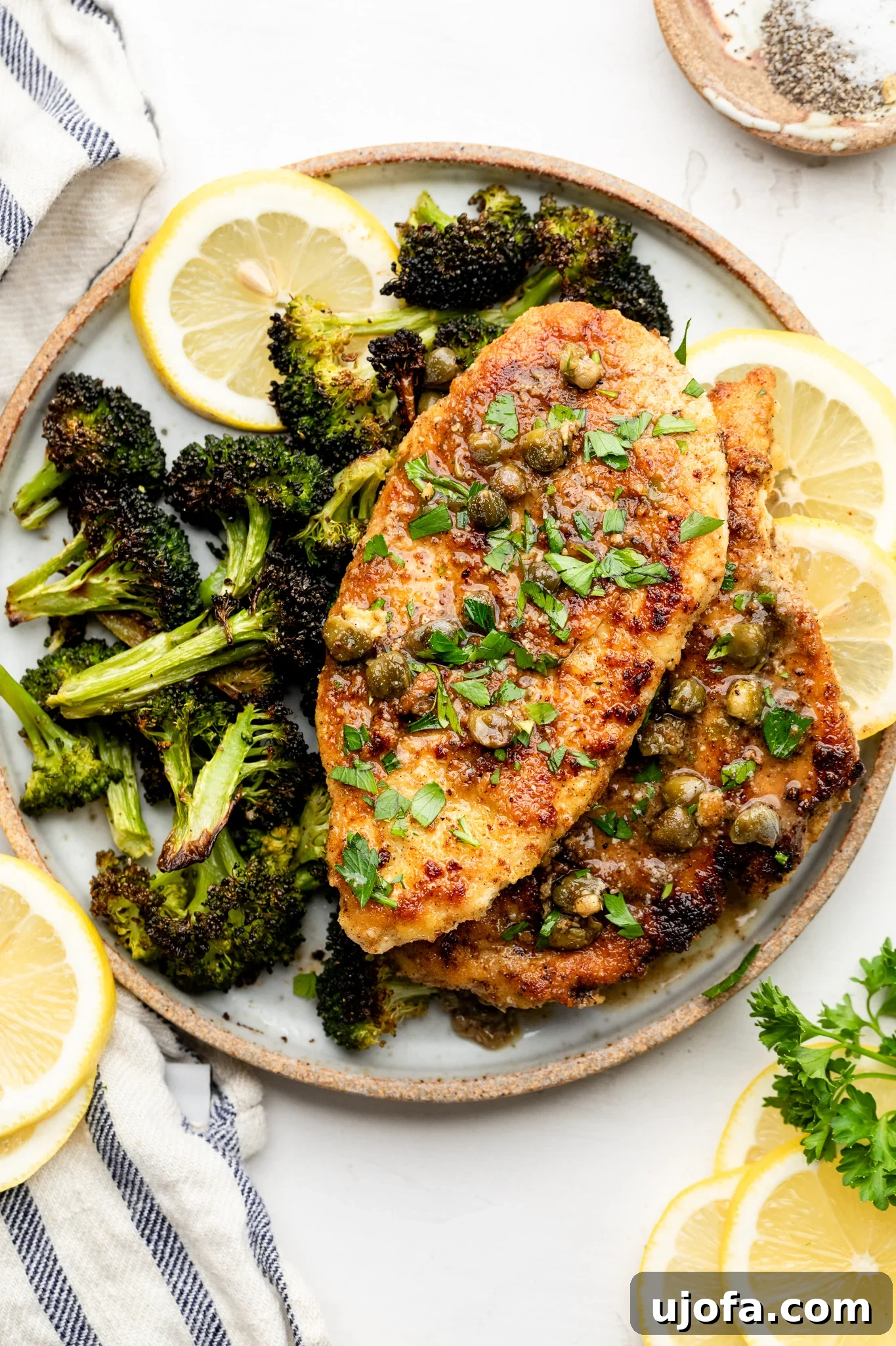 A tan and white plate with roasted broccoli, gluten free chicken piccata, and lemon slices