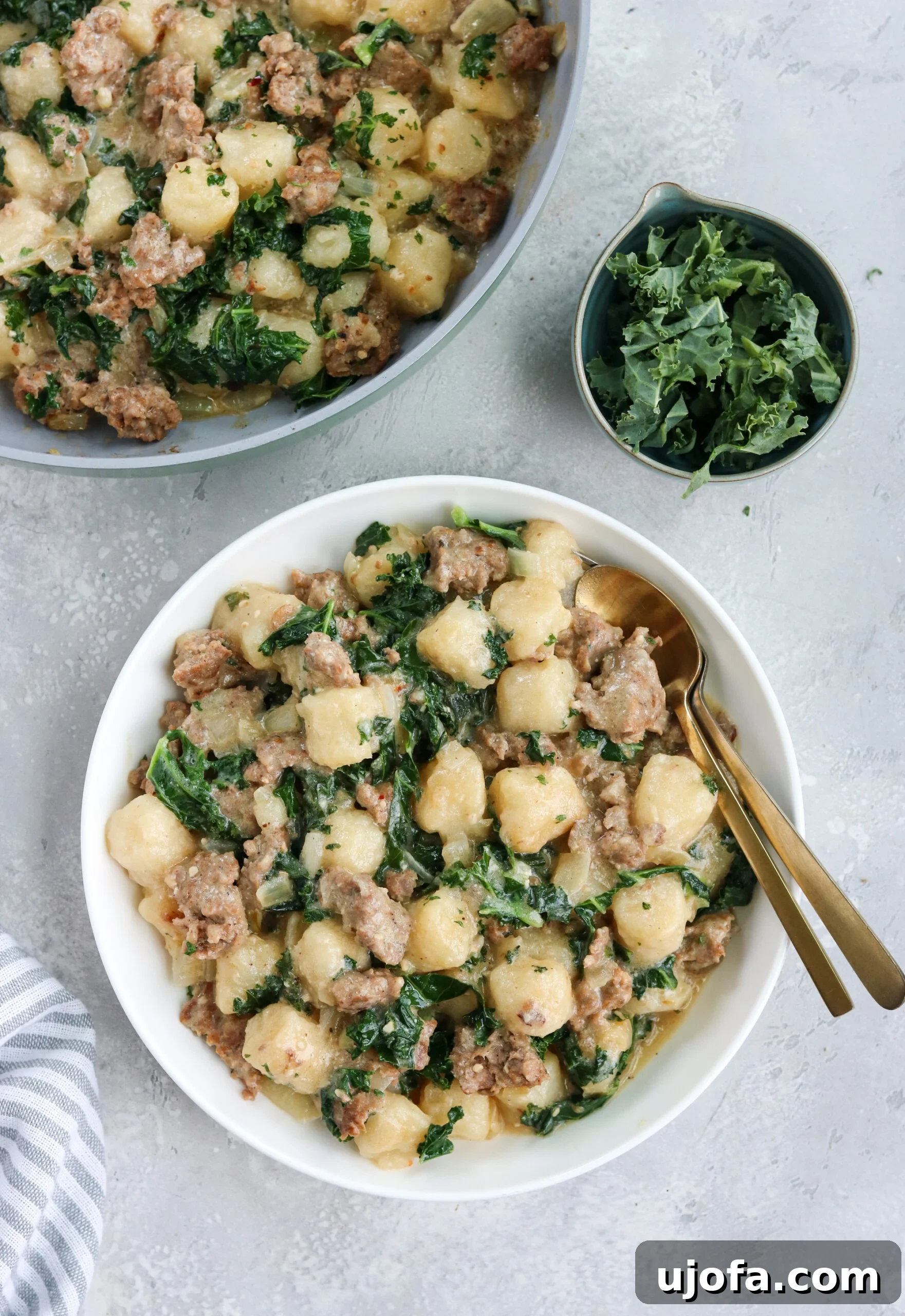 A vibrant bowl of Paleo Sausage Kale Gnocchi, featuring golden-brown gnocchi, savory sausage, and wilted kale, garnished and ready to serve. Two gold spoons rest beside it, with a bowl of fresh kale and a skillet of gnocchi in the background, all on a clean white surface.