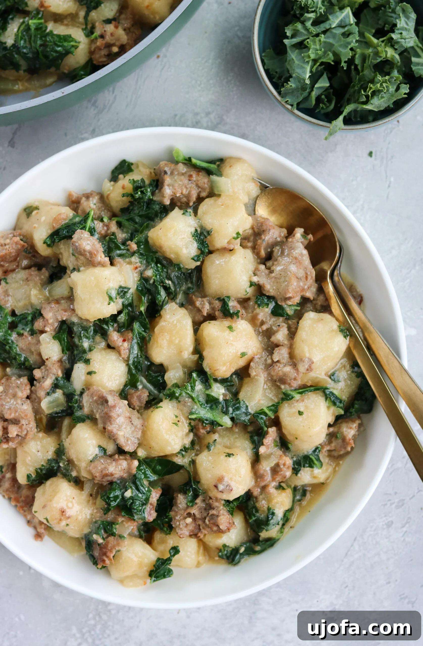 Close-up of a bowl of Paleo Sausage Kale Gnocchi, showcasing the crispy cauliflower gnocchi, chunks of Italian sausage, and tender kale, with a skillet of the dish in the background and a fresh bowl of kale, all on a pristine white surface.
