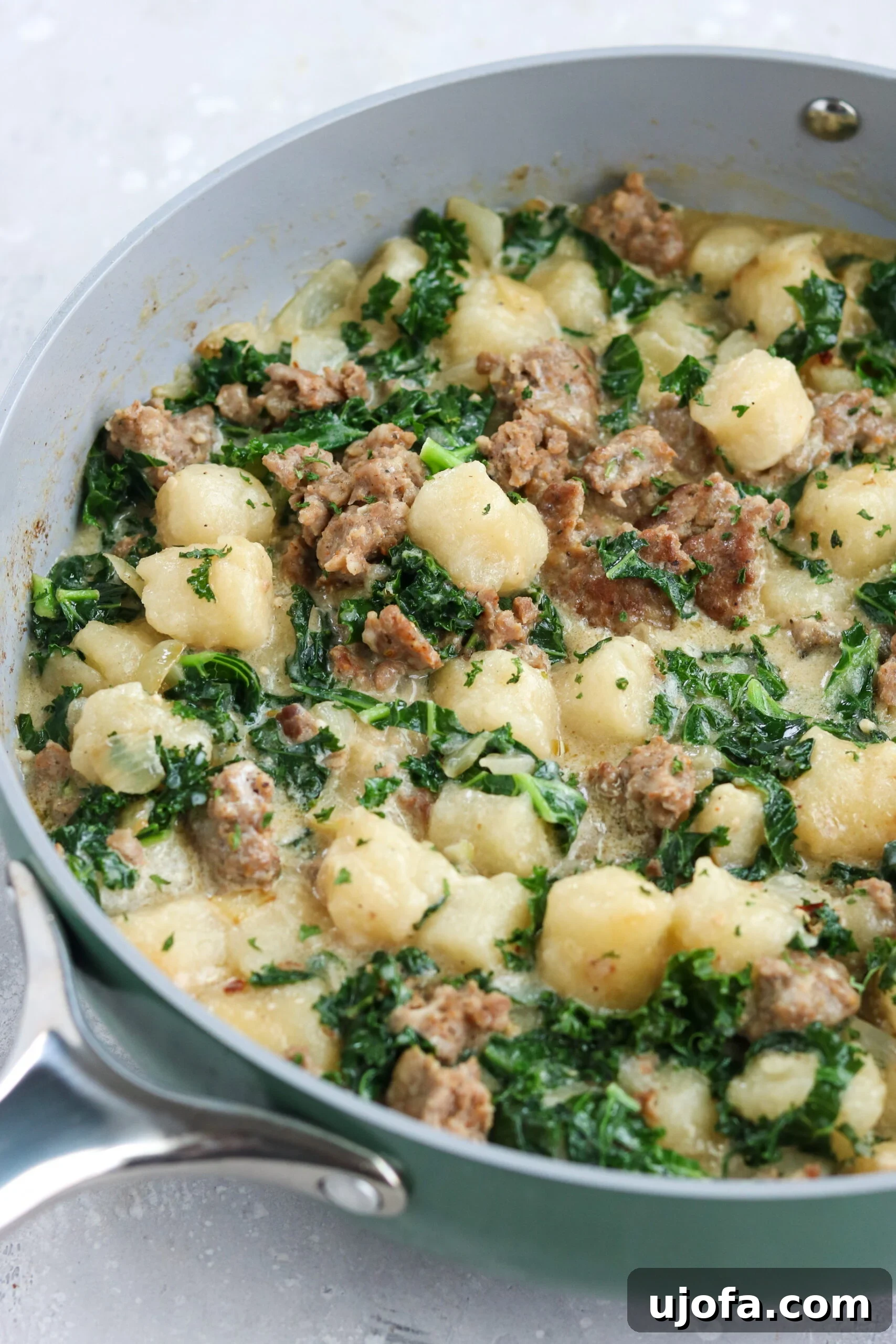 A close-up of Paleo Sausage Kale Gnocchi cooking in a sleek gray non-stick skillet with a silver handle, highlighting the perfectly browned gnocchi and tender kale, all on a minimalist white background.