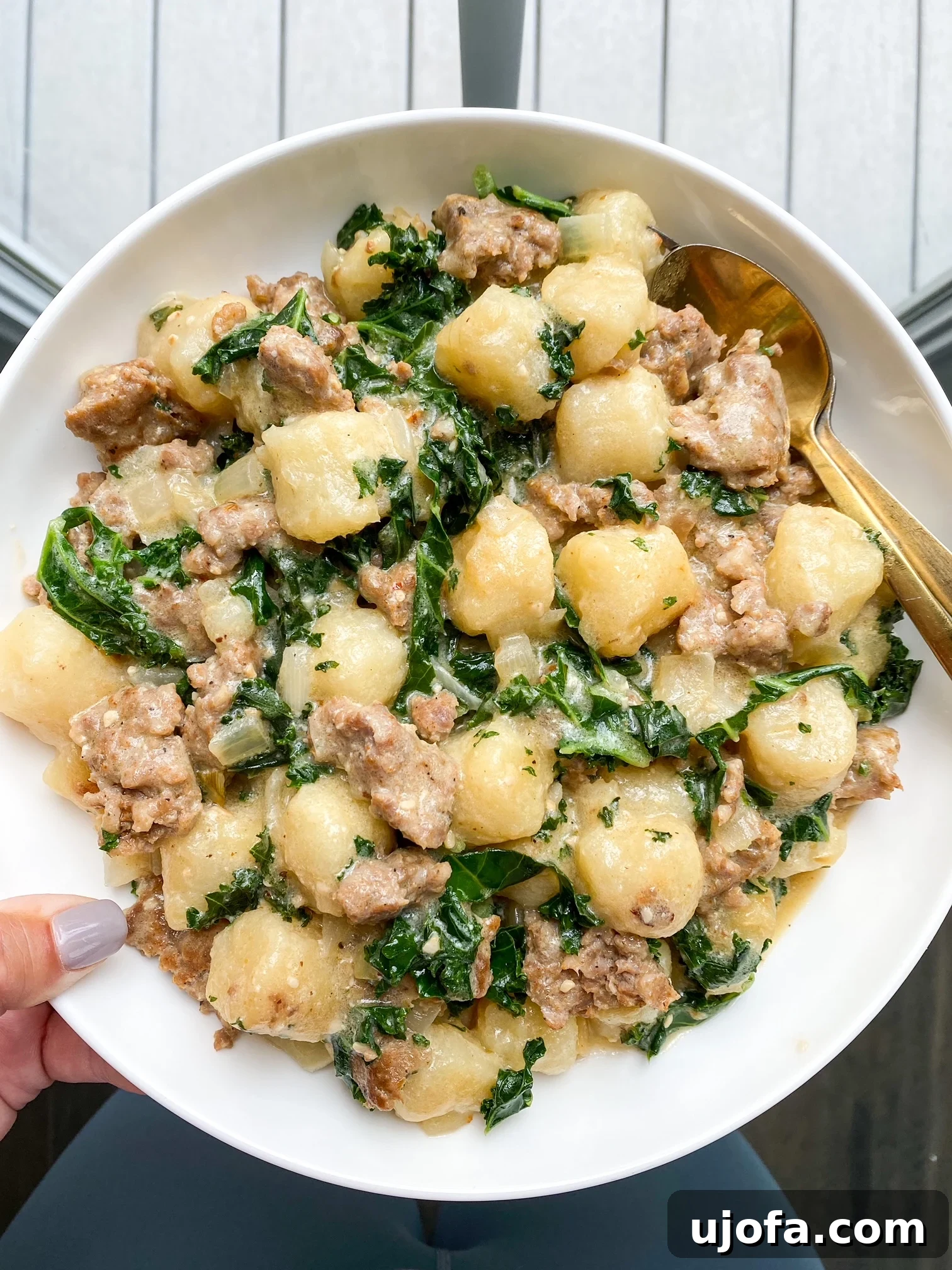 A person holding a white bowl filled with a generous serving of Paleo Sausage Kale Gnocchi, showcasing the rich texture and inviting colors of the dish, ready to be enjoyed.