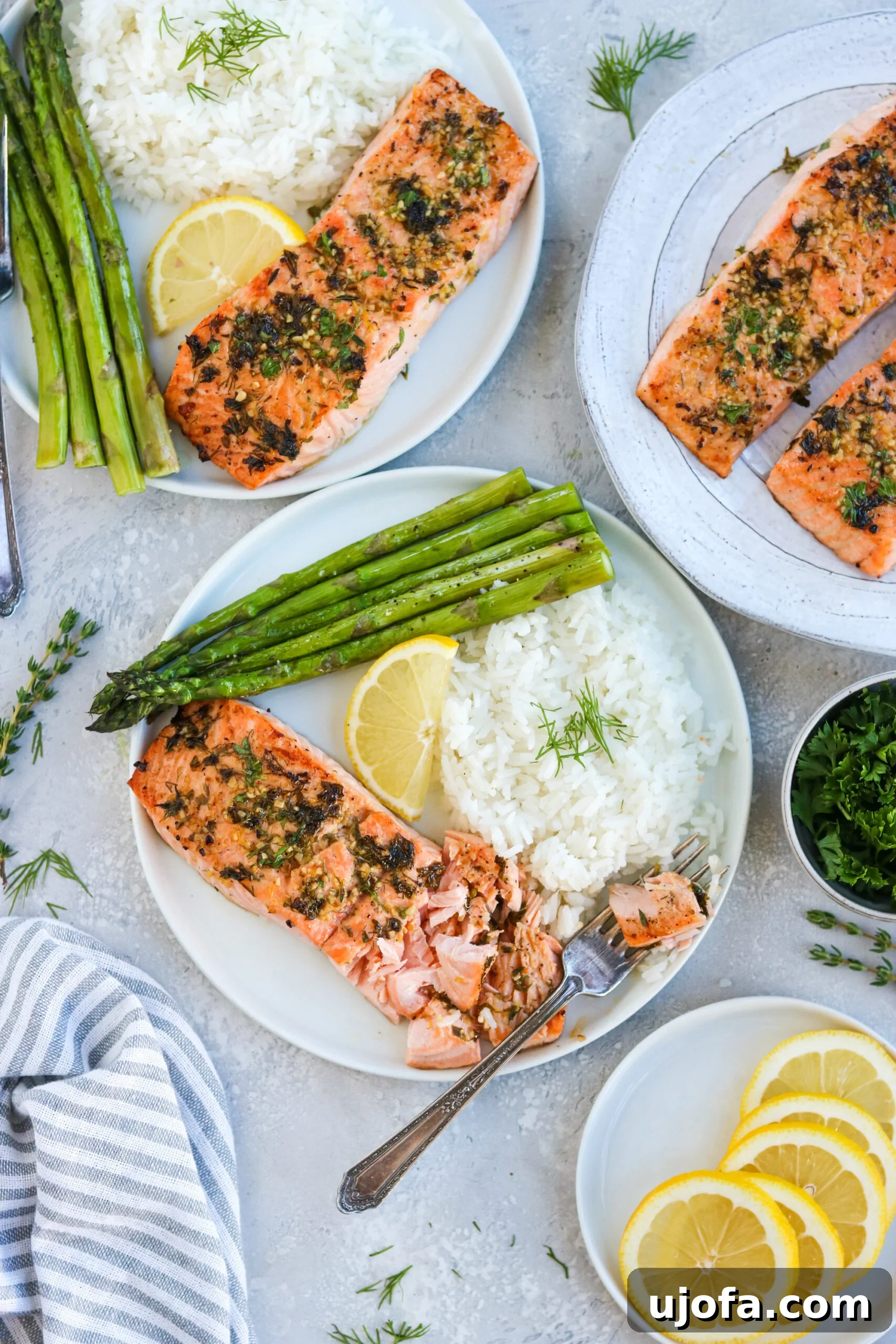 Multiple Servings of Garlic Herb Baked Salmon Three servings of garlic herb baked salmon on white plates, each accompanied by asparagus, white rice, and a lemon wedge, illustrating perfect plating.