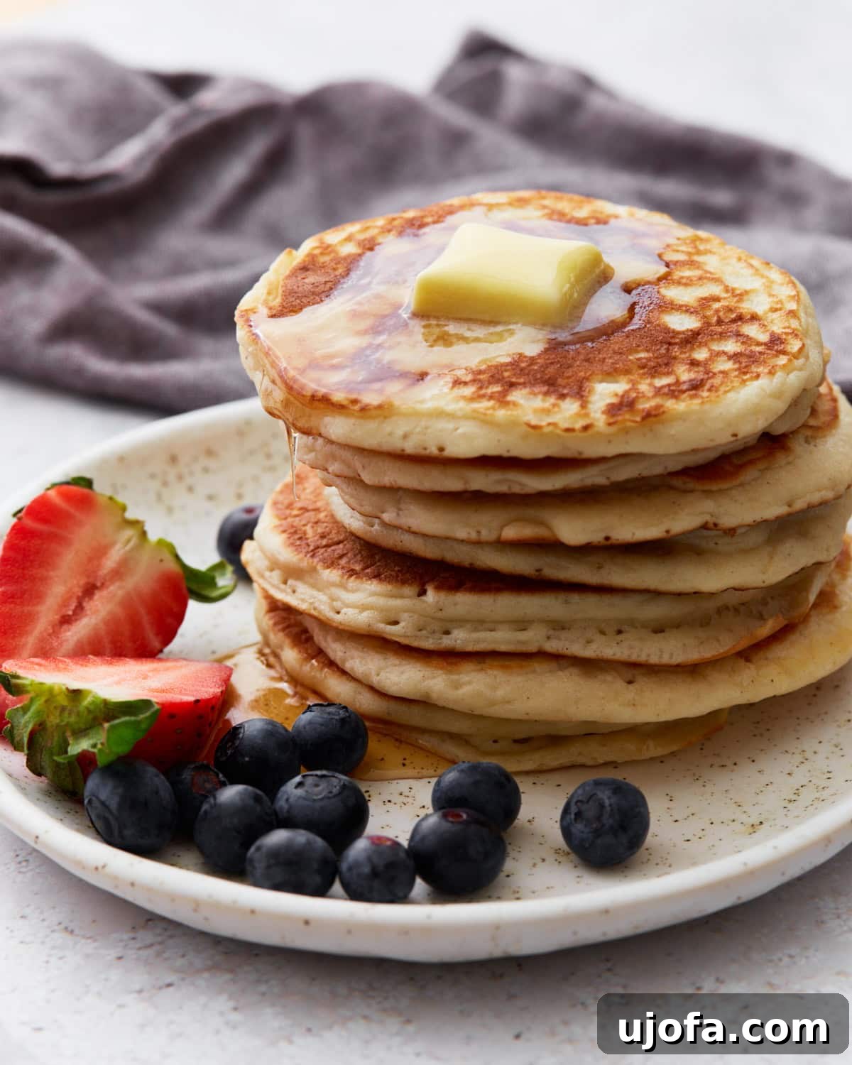 Foolproof Gluten-Free Pancakes 2 A stack of fluffy gluten-free pancakes topped with melting butter and drizzled with syrup, served on a speckled white plate with fresh strawberries and blueberries on the side. A dark napkin is in the background.