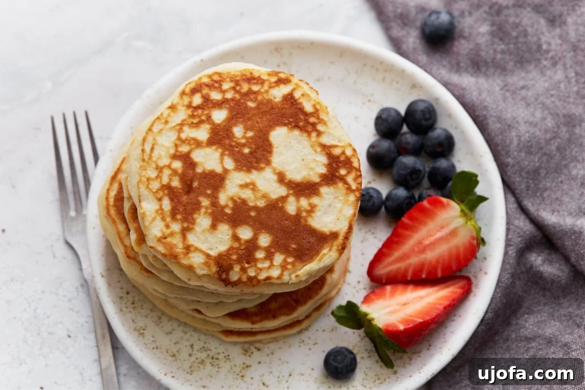 Foolproof Gluten-Free Pancakes 11 A stack of golden gluten-free pancakes on a white speckled plate, garnished with fresh blueberries and sliced strawberries, with a fork placed beside the plate.