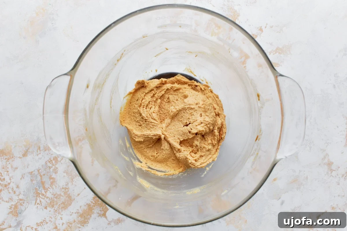 Glass mixing bowl with creamy peanut butter, sugars, and butter, ready for blending on a light textured surface.