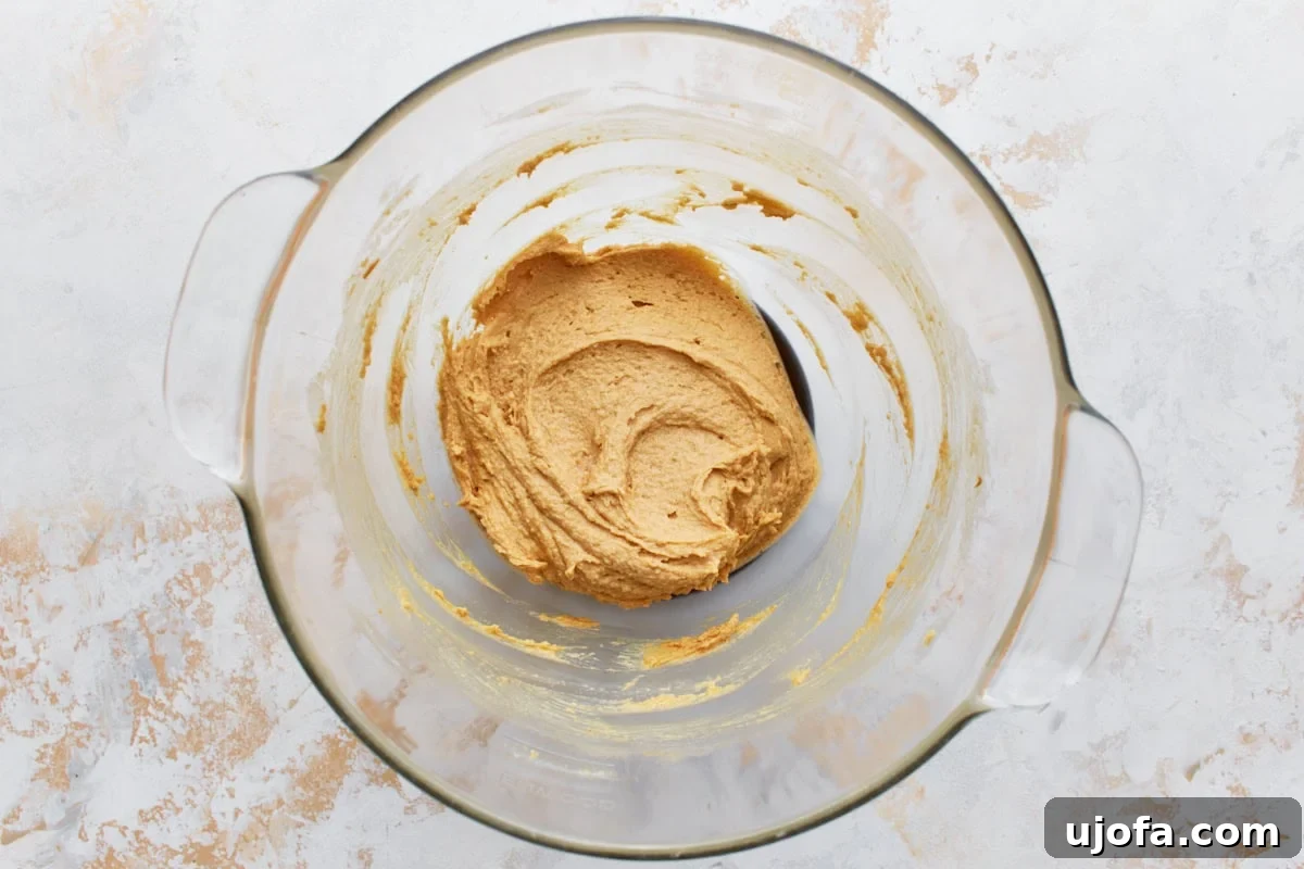 Glass mixing bowl with creamy peanut butter mixture and a single egg, ready to be incorporated on a light textured surface.