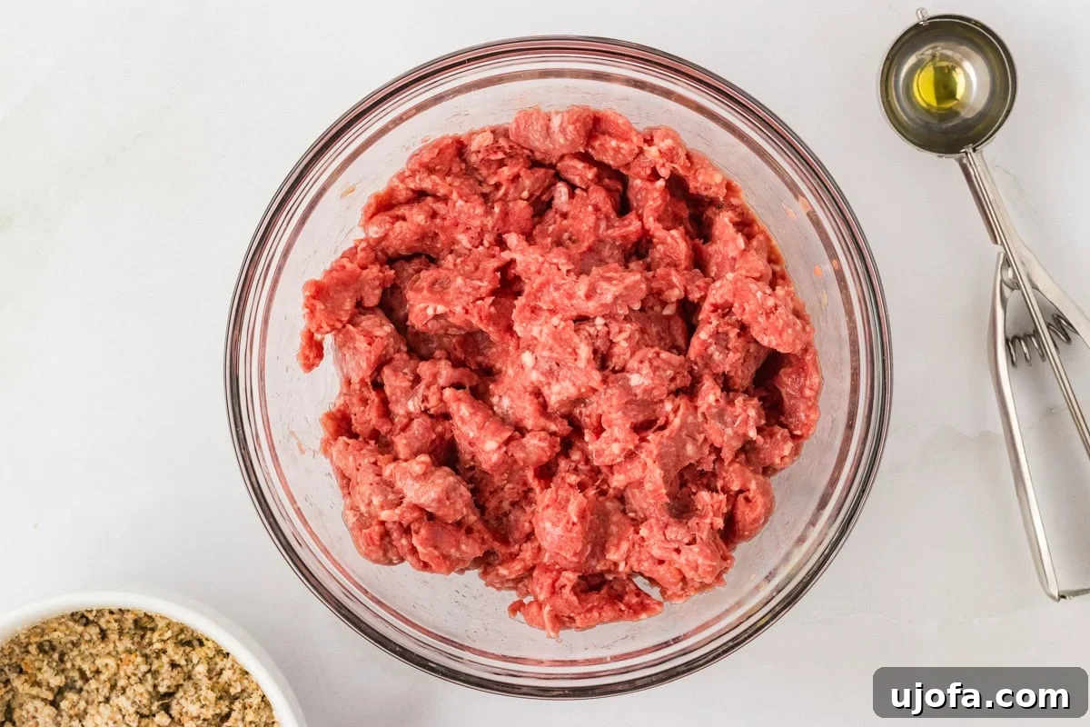 Raw ground beef in a glass mixing bowl with a cookie scoop and measuring spoon beside it.