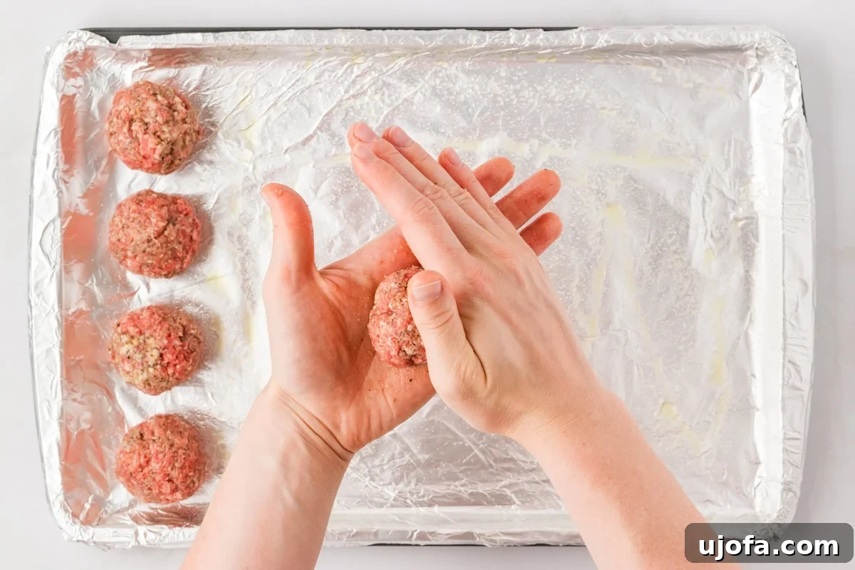 Hands shaping a meatball over a foil-lined baking sheet with already formed meatballs in the background.