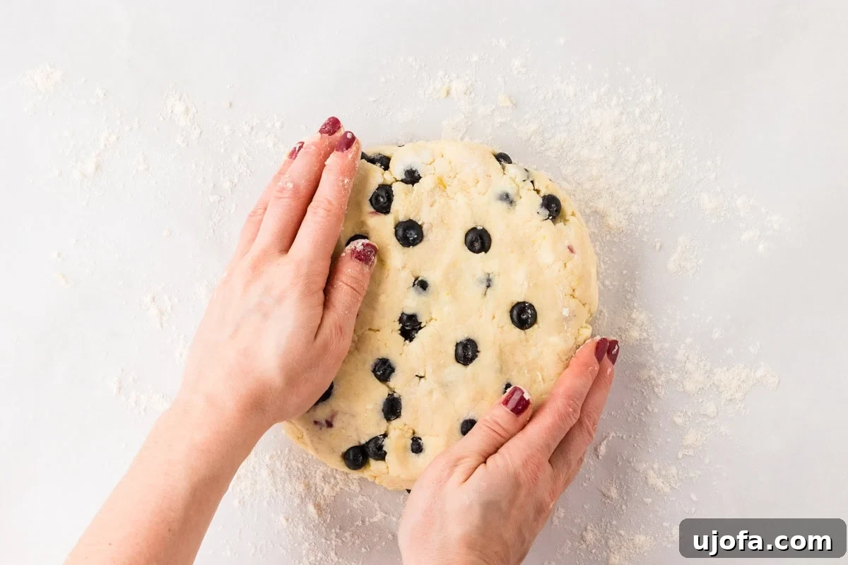Blueberry Burst Gluten-Free Scones 13 Hands shaping the gluten-free blueberry scone dough into a flat round disk on a floured surface, with blueberries visible in the dough.