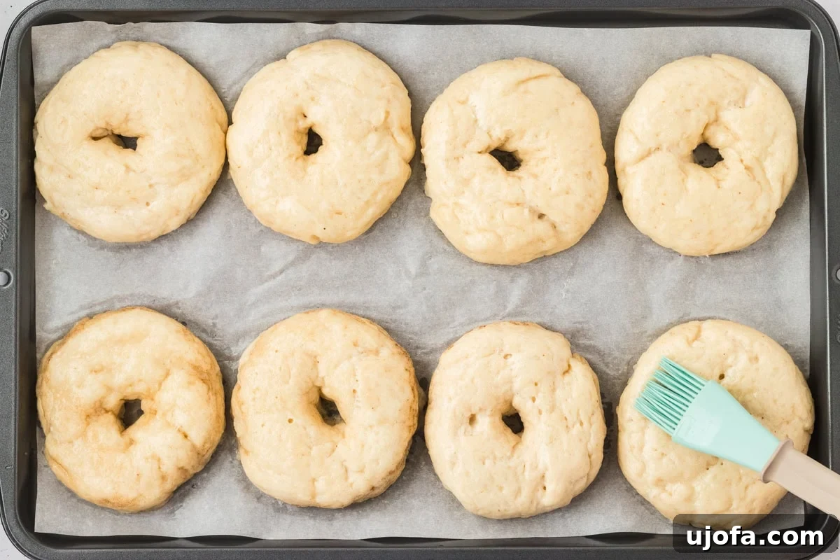 Par-boiled gluten-free bagels on a baking tray being brushed with egg wash before baking.
